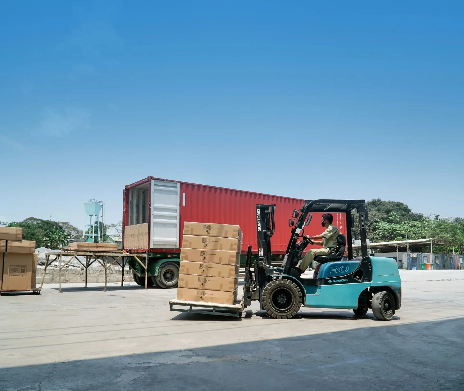 A forklift carrying a pallet of cartons toward an open shipping container in a loading yard.
